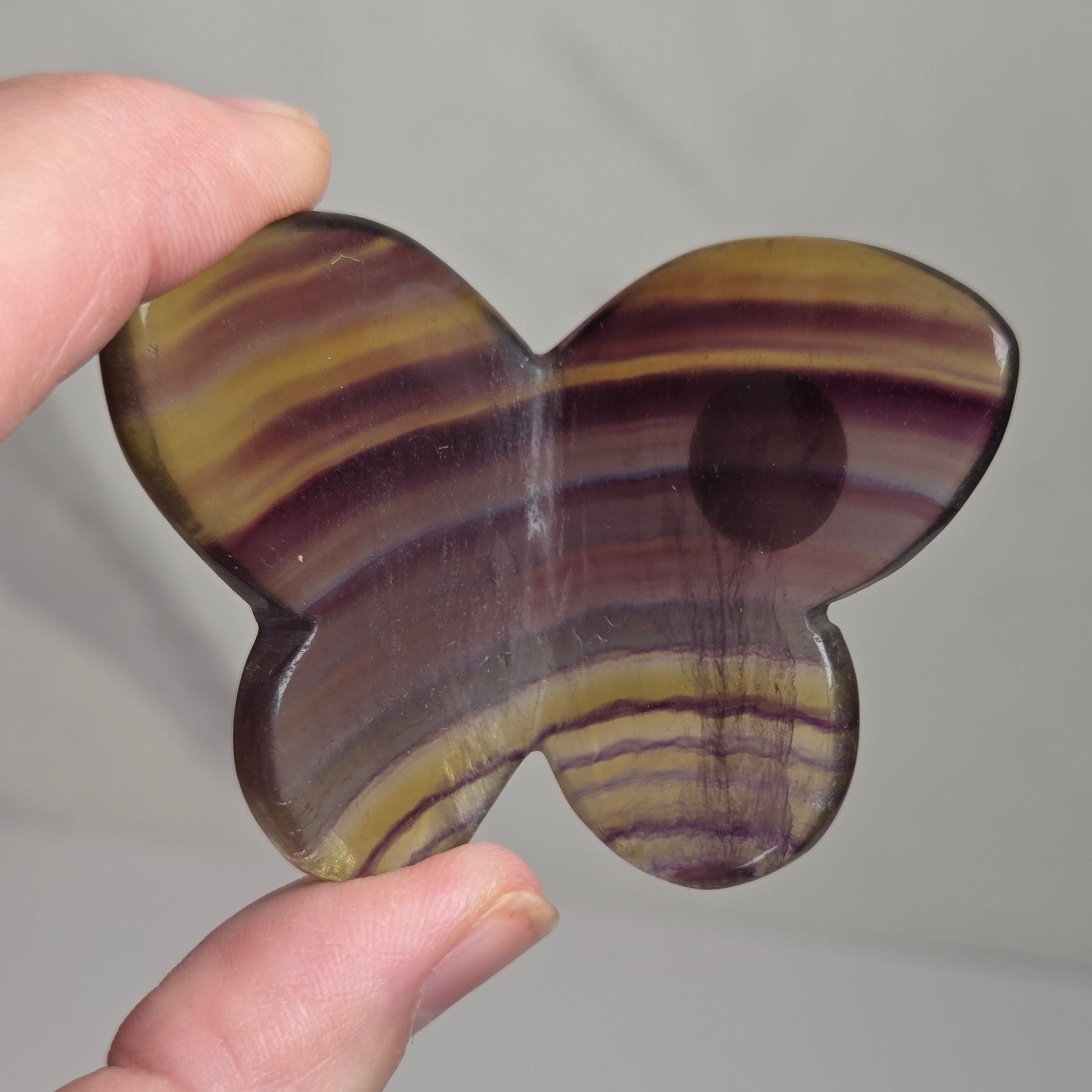 A hand holding a butterfly-shaped polished stone piece with striped bands of purple, brown, and yellow, against a light gray background.