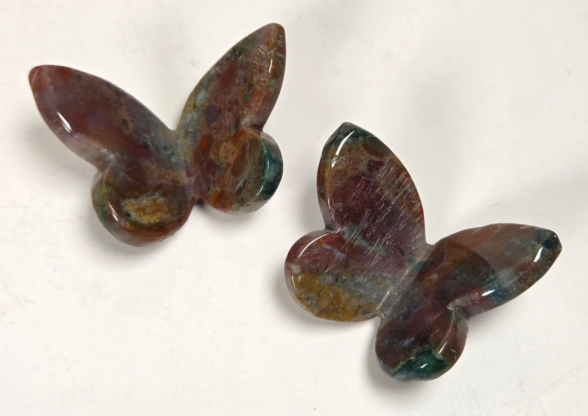 Two polished butterfly-shaped stone carvings with a mottled brown, green, and purple patina, displayed on a plain off-white surface.