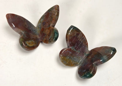 Two polished butterfly-shaped stone carvings with a mottled brown, green, and purple patina, displayed on a plain off-white surface.