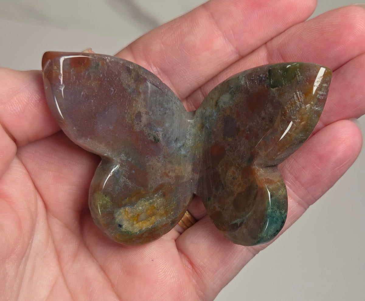 A hand holding a polished butterfly-shaped stone with mottled brown, green, and gray tones.
