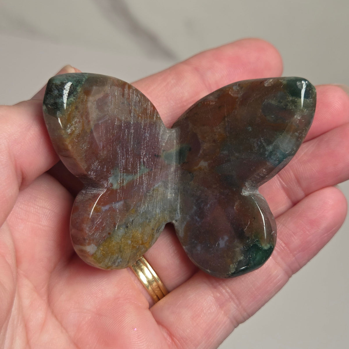 A hand holding a polished butterfly-shaped stone with mottled brown, green, and purple tones, with a gold ring on one finger, against a light gray marble-patterned background.