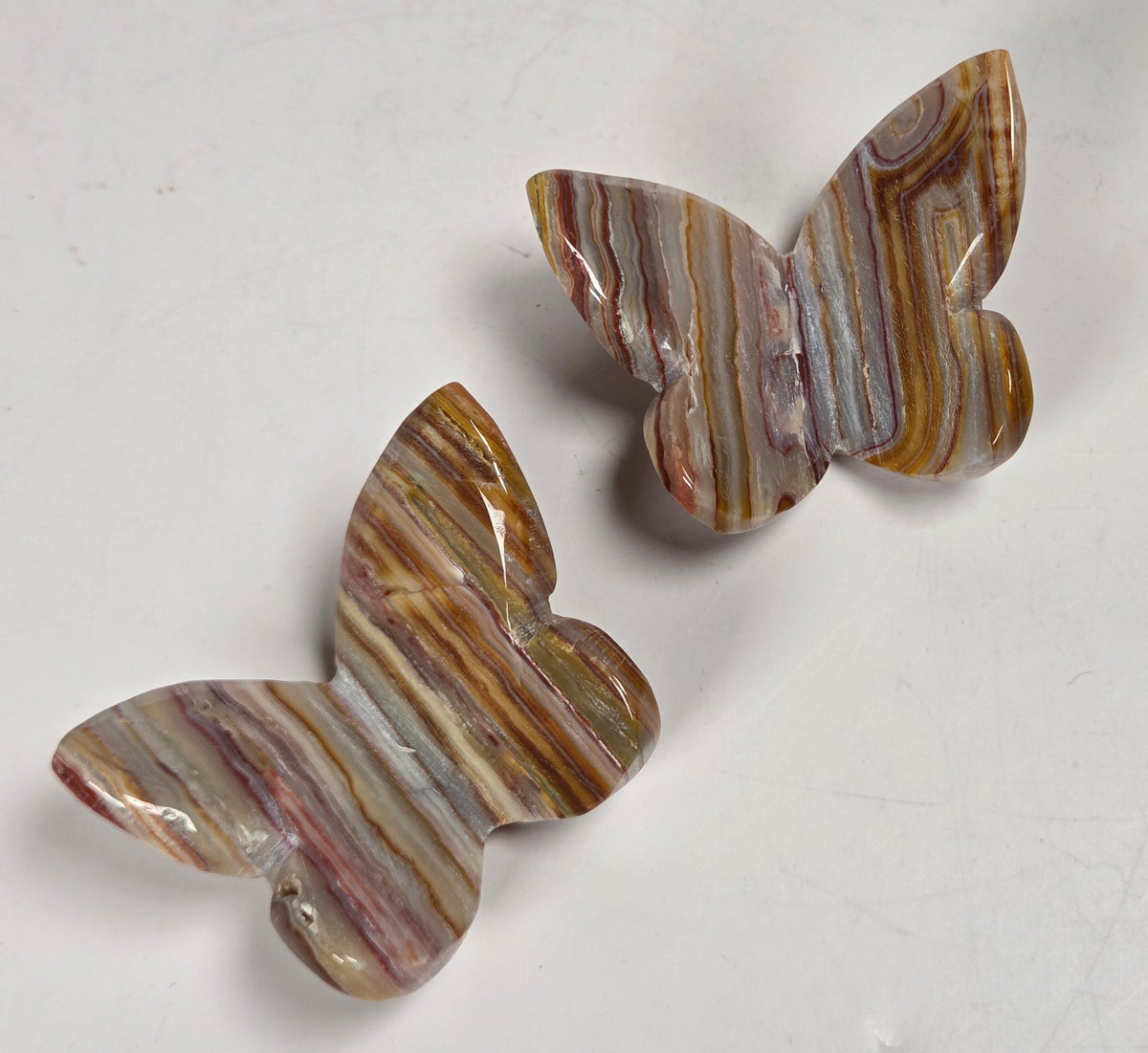 Two butterfly-shaped stone jewelry pieces with natural striped patterns in brown, beige, and muted pink, displayed on a light gray surface.