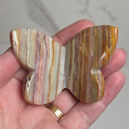 A hand holding a polished butterfly-shaped stone piece with striped patterns in brown, gold, beige, and white, set against a light gray marble background.
