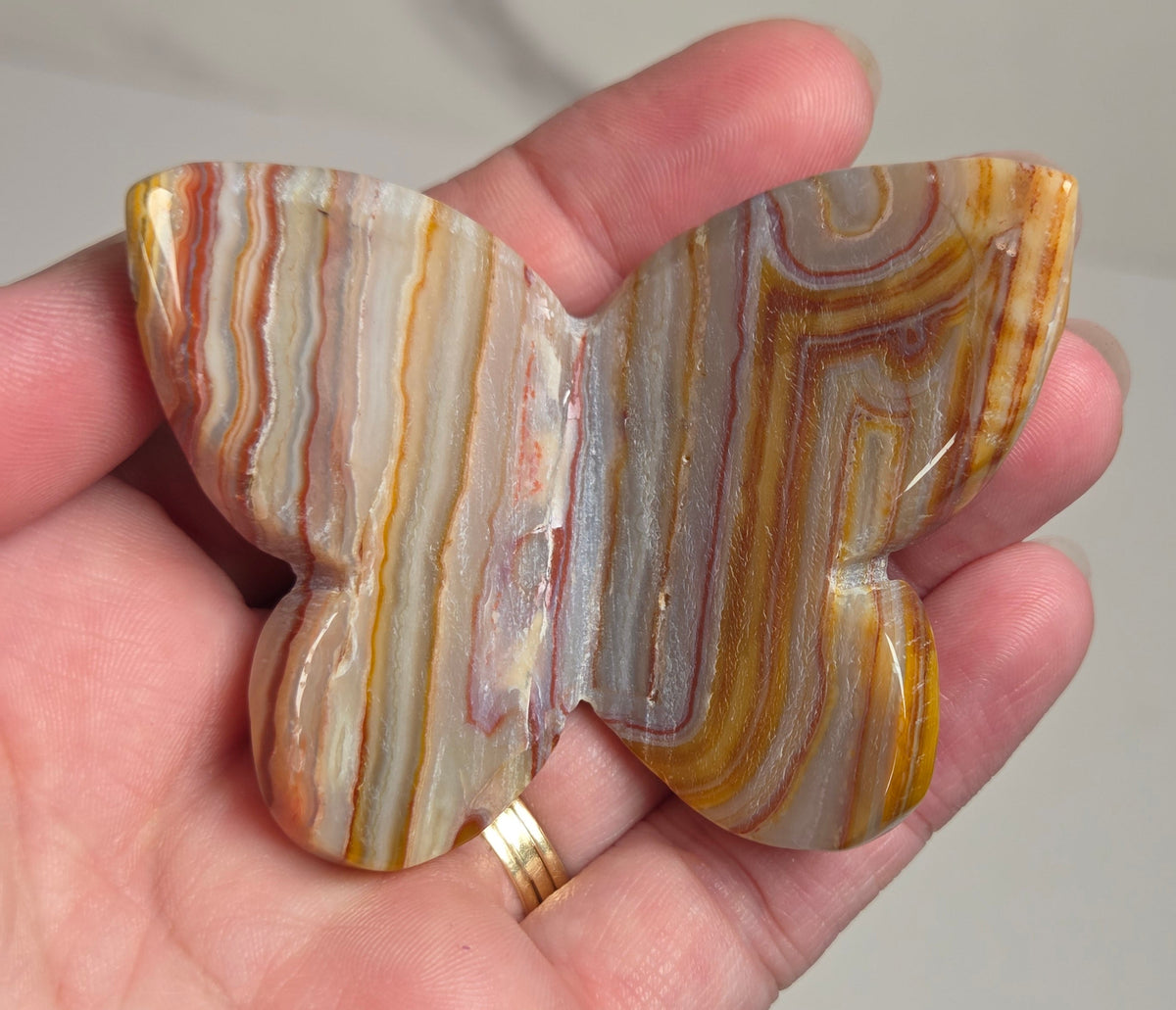 A hand holding a butterfly-shaped polished stone ornament with natural agate-like banding in cream, brown, orange, and gray tones.