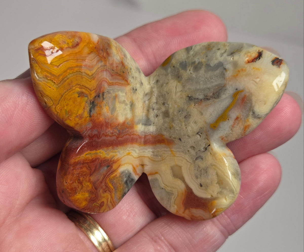 A hand holding a polished butterfly-shaped stone carving with marbled orange, yellow, brown, and gray patterns.