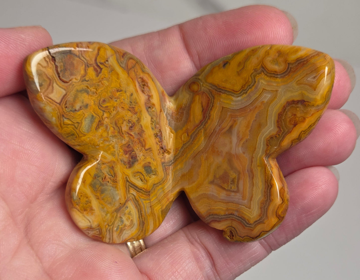 A hand holding a polished butterfly-shaped stone pendant or brooch with swirling golden yellow, orange, and brown agate-like patterns.