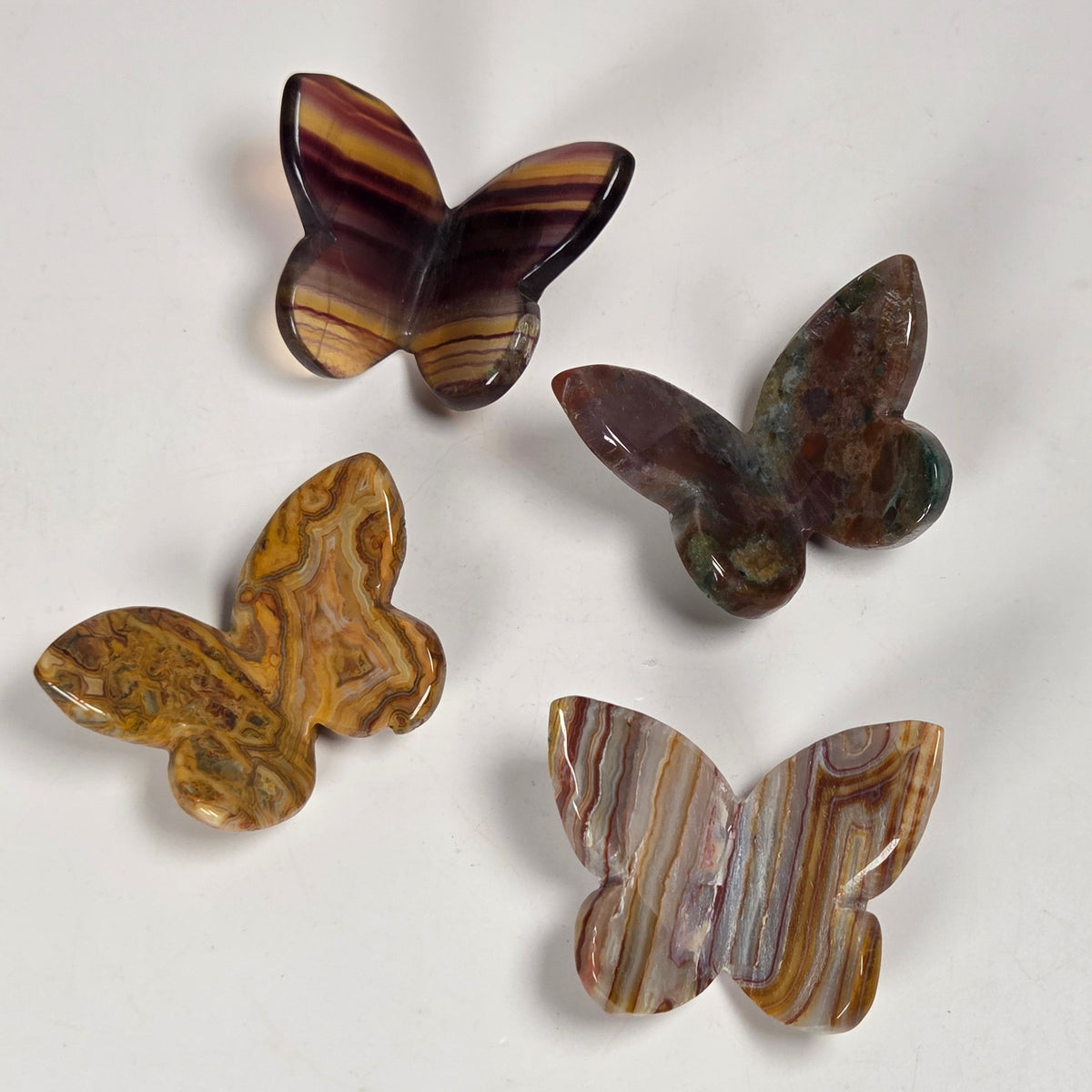 Four carved butterfly-shaped stone pieces with natural striped patterns in brown, purple, and gold tones, arranged on a plain off-white surface.