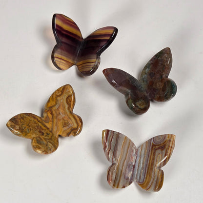Four carved butterfly-shaped stone pieces with natural striped patterns in brown, purple, and gold tones, arranged on a plain off-white surface.