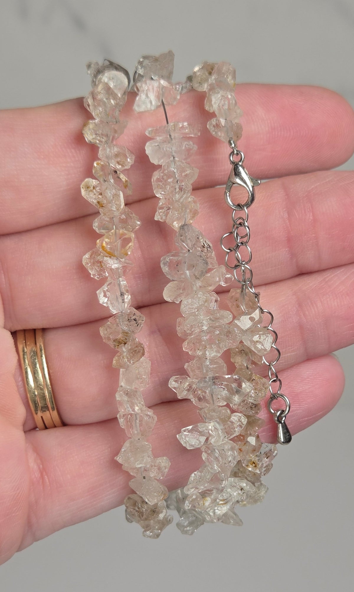 A close-up of a hand holding two beaded crystal bracelets. The bracelets are made of irregular, translucent clear quartz chips with some warm yellowish tones, and one has a silver clasp with an extender chain.