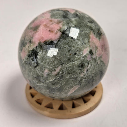 A polished pink and green stone sphere resting on a small round wooden display stand against a plain light background.