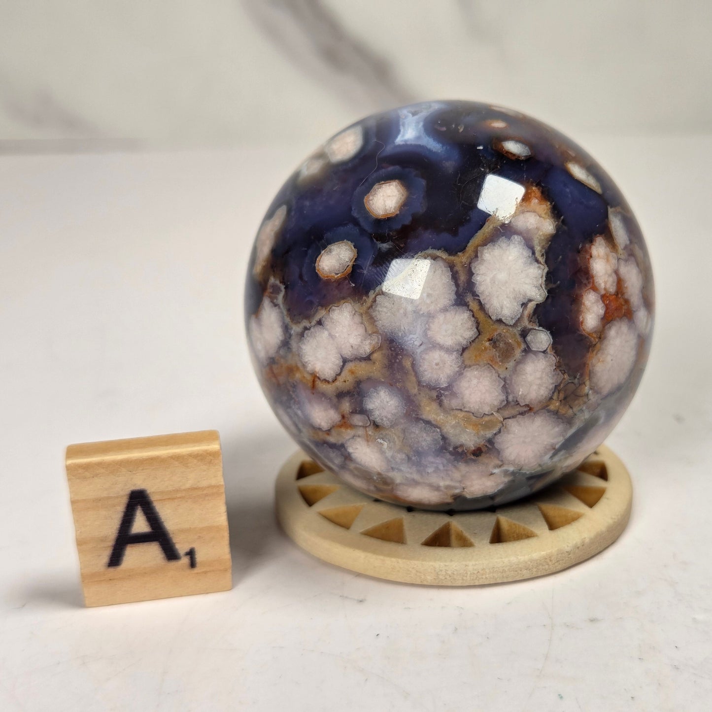 A polished agate crystal sphere with a blue, white, and brown marbled pattern, displayed on a small wooden stand next to a wooden letter tile showing the letter 'A'.