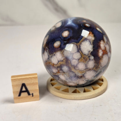 A polished agate crystal sphere with a blue, white, and brown marbled pattern, displayed on a small wooden stand next to a wooden letter tile showing the letter 'A'.