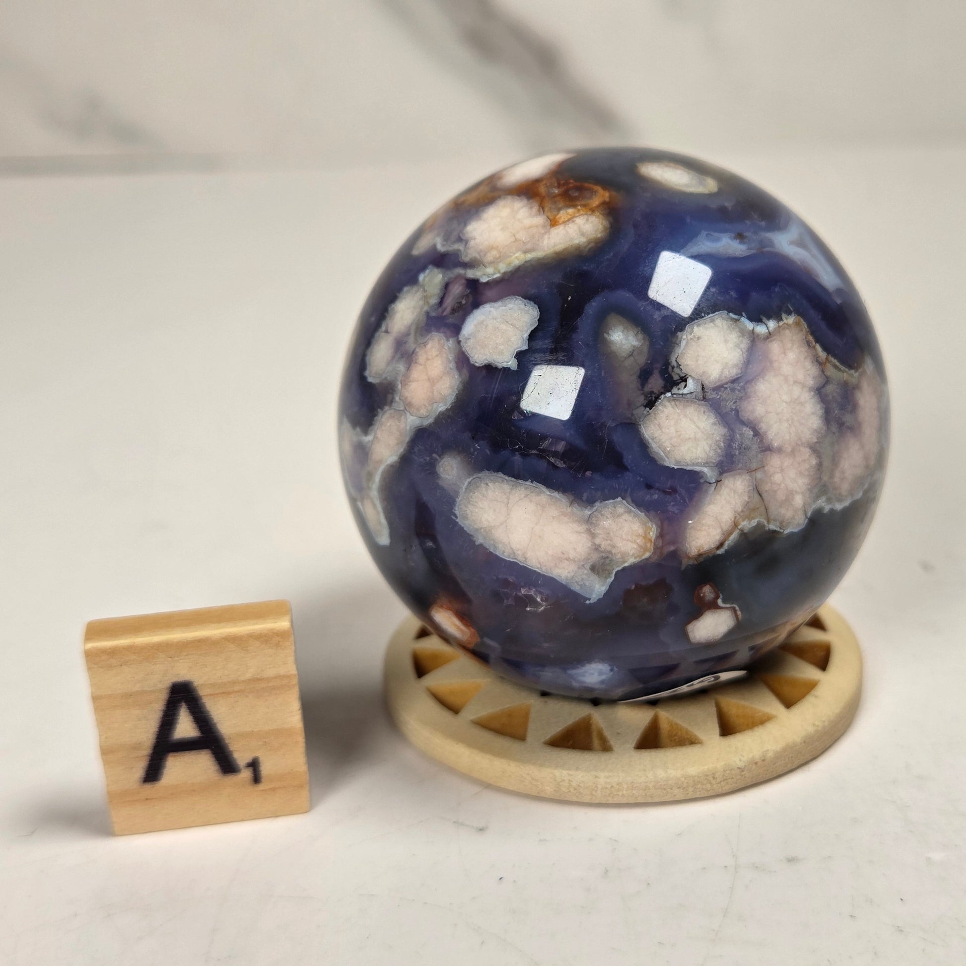 A polished agate crystal sphere with deep blue, white, and brown natural patterns, displayed on a small wooden stand next to a wooden letter tile spelling 'A1' on a light surface.