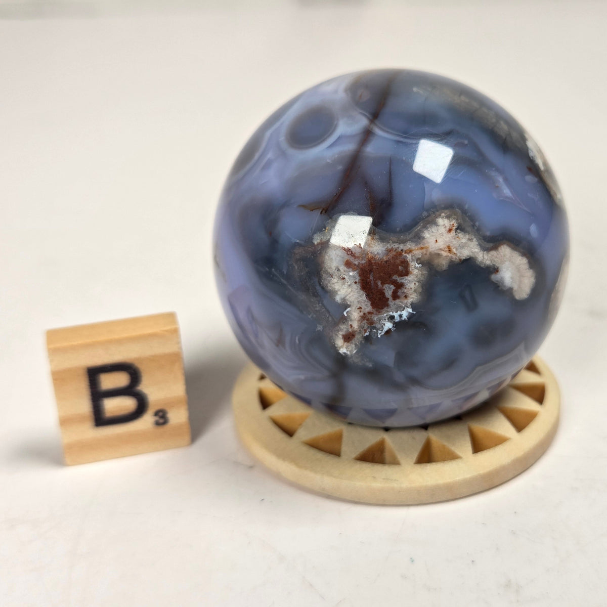 A polished blue agate crystal sphere with natural brown and white matrix patterns, displayed on a light wood circular stand next to a wooden Scrabble tile showing the letter B.