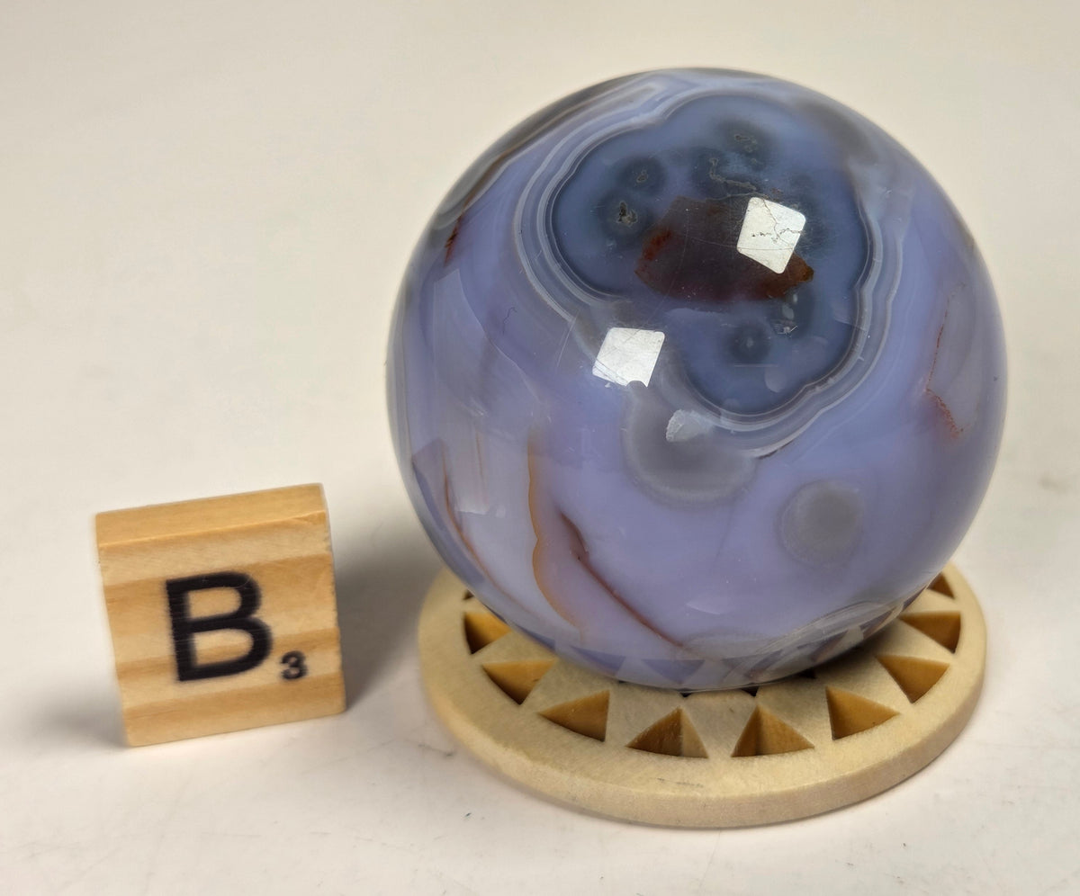A polished agate crystal sphere with swirling lavender, blue, and white banding sits on a small carved wooden stand. A wooden letter tile showing the letter 'B' is placed beside it on a light neutral surface.