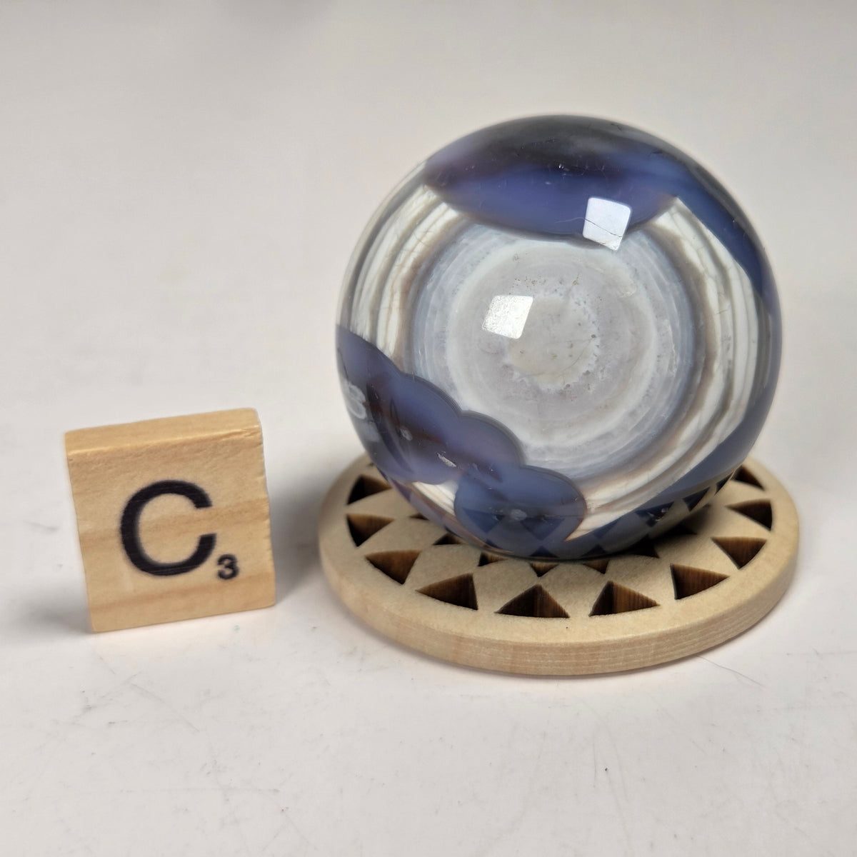 A round glass decorative object with swirling blue, white, and gray marbled patterns, displayed on a small round wooden stand next to a Scrabble tile showing the letter C.