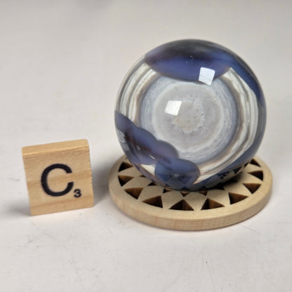 A round glass decorative object with swirling blue, white, and gray marbled patterns, displayed on a small round wooden stand next to a Scrabble tile showing the letter C.