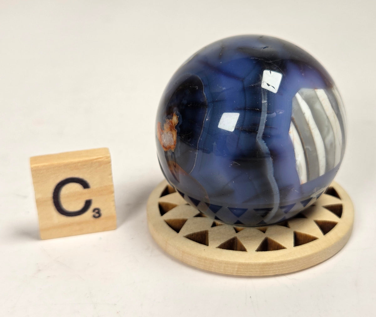A polished blue agate crystal sphere resting on a circular wooden stand, with a small wooden Scrabble tile displaying 'C3' to the left, set against a plain off-white background.