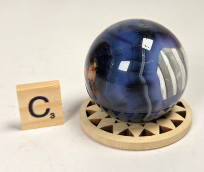 A polished blue agate crystal sphere resting on a circular wooden stand, with a small wooden Scrabble tile displaying 'C3' to the left, set against a plain off-white background.