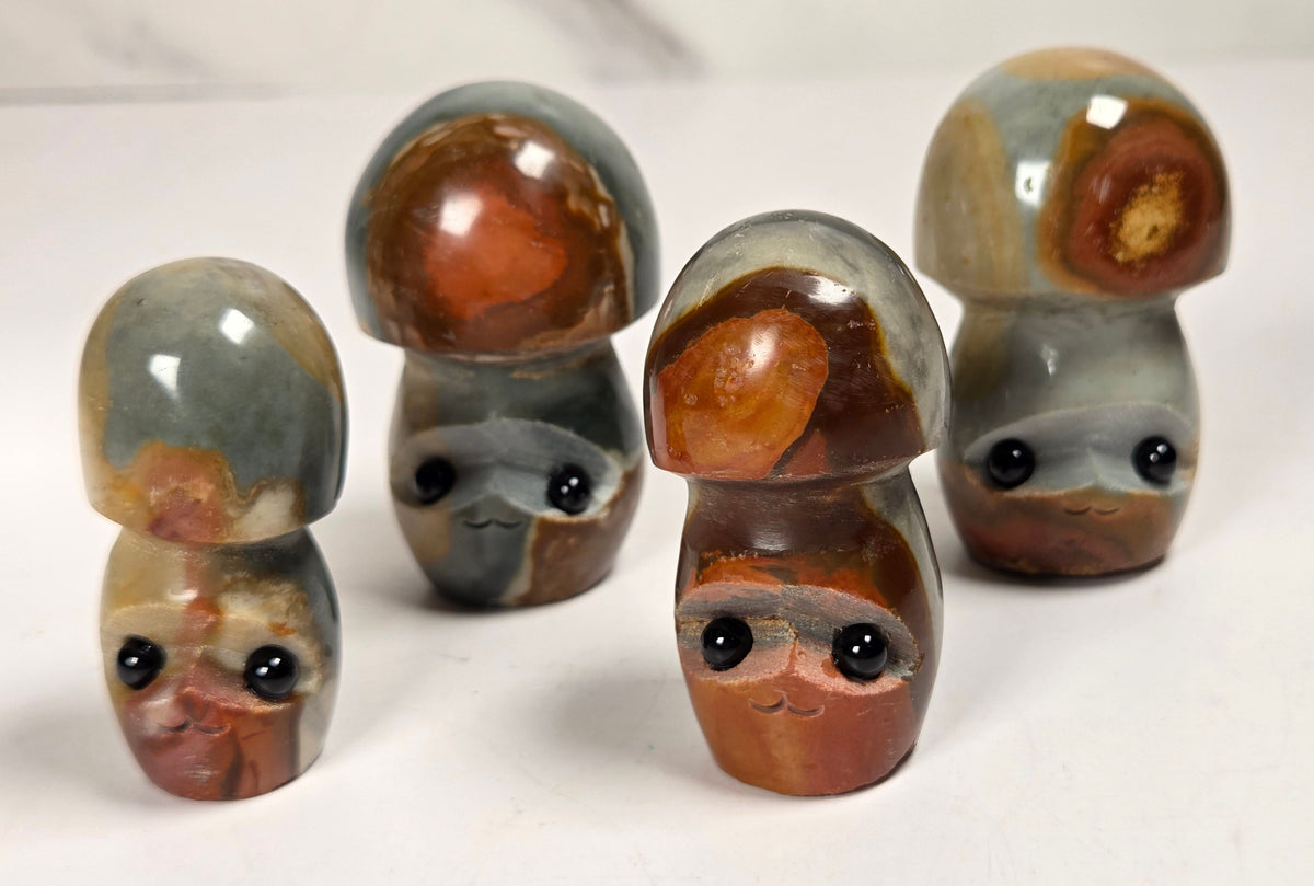 Four small mushroom-shaped figurines carved from agate, featuring natural banded patterns in gray, brown, and rust orange, with cute black bead eyes and simple carved facial features, arranged on a white surface.