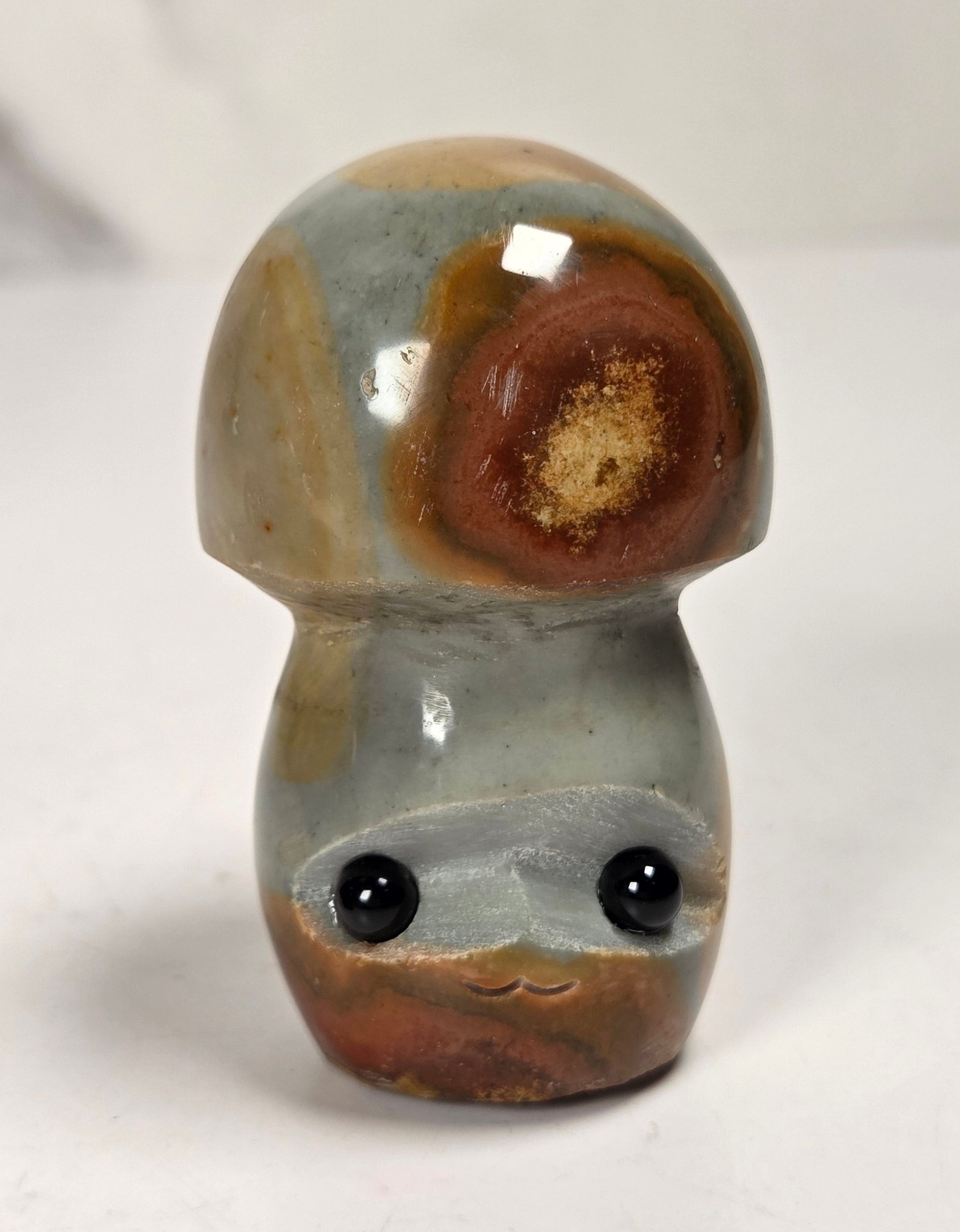A small mushroom-shaped decorative object with a glossy marbled finish in gray, brown, and tan, featuring two black bead eyes, photographed on a light neutral surface.