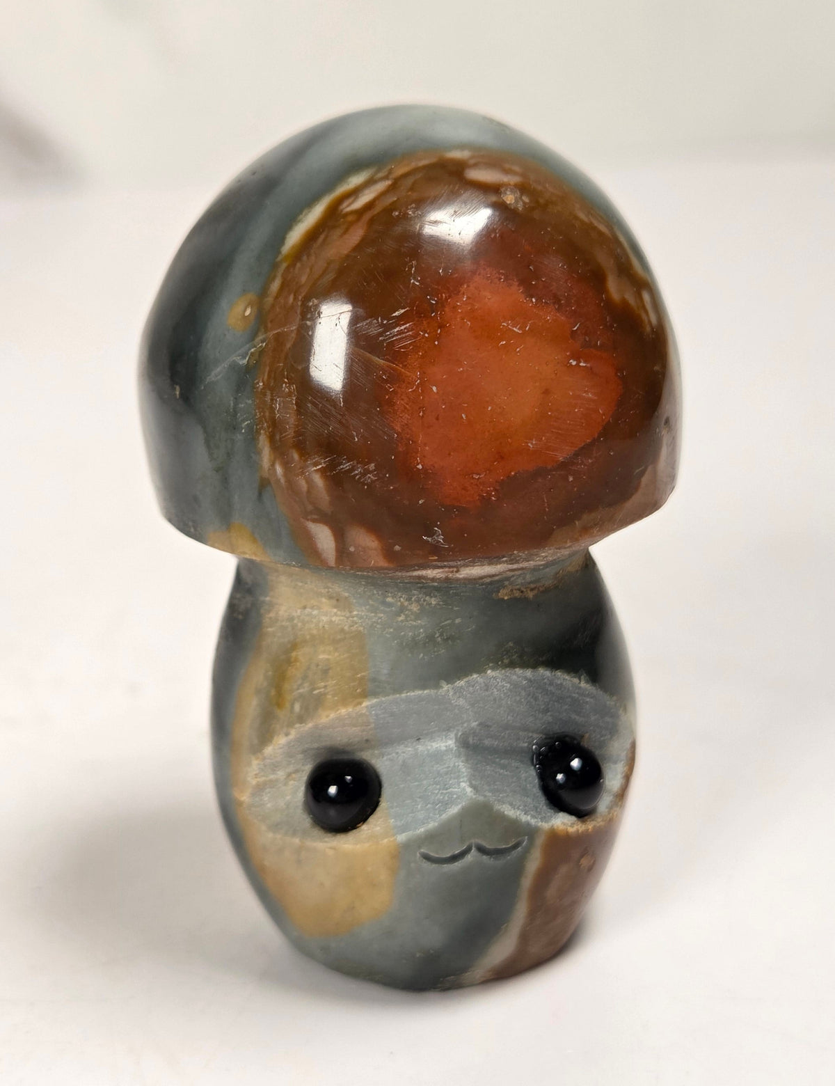 A small mushroom-shaped figurine carved from polished agate, featuring a glossy reddish-brown cap and a mottled gray, brown, and beige base with black bead eyes and a stitched smile, set against a plain light background.
