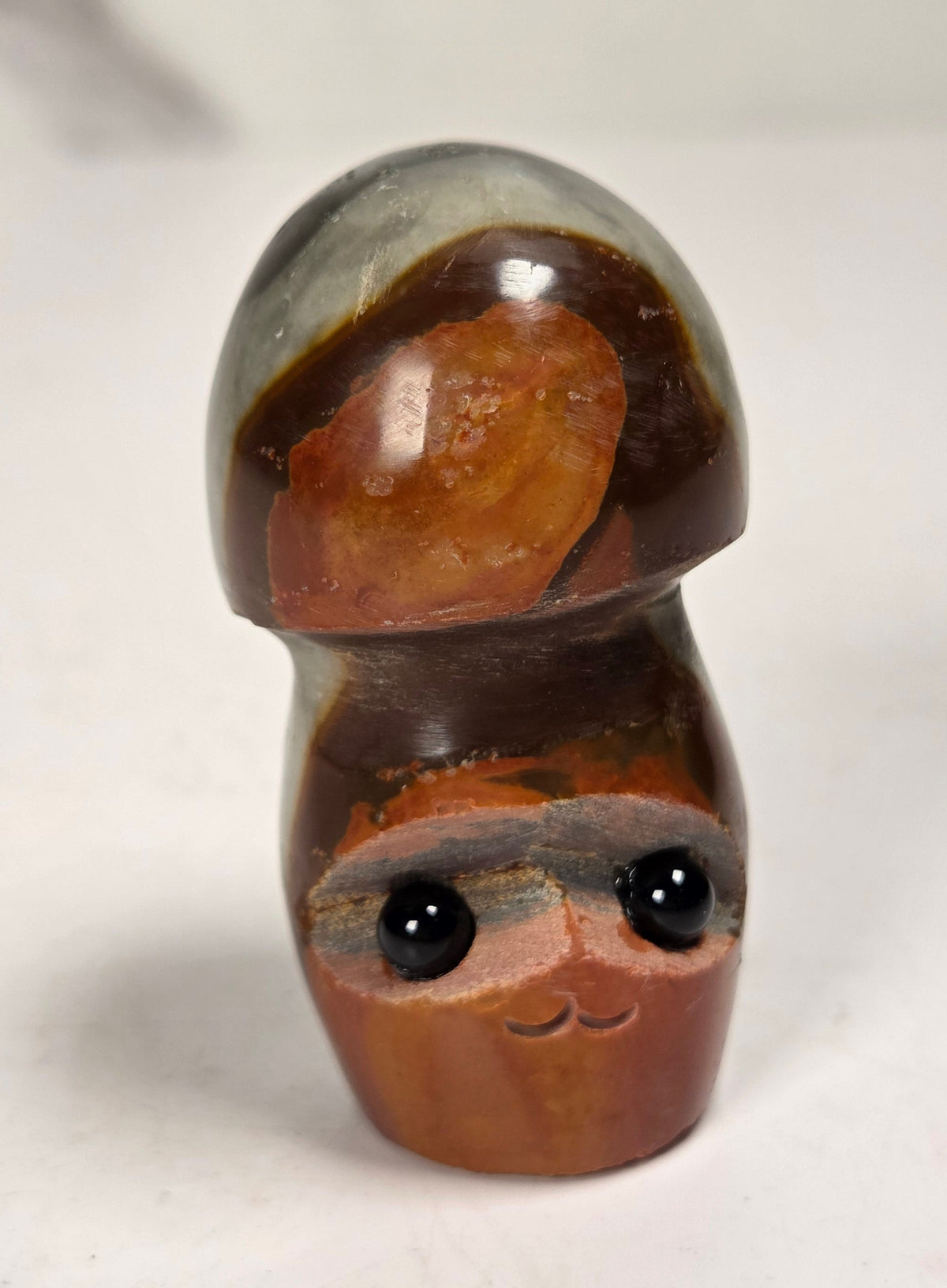 A small carved wooden figurine with a mushroom cap on top and a smiling face on the stem, featuring glossy black bead eyes, set against a plain light background.