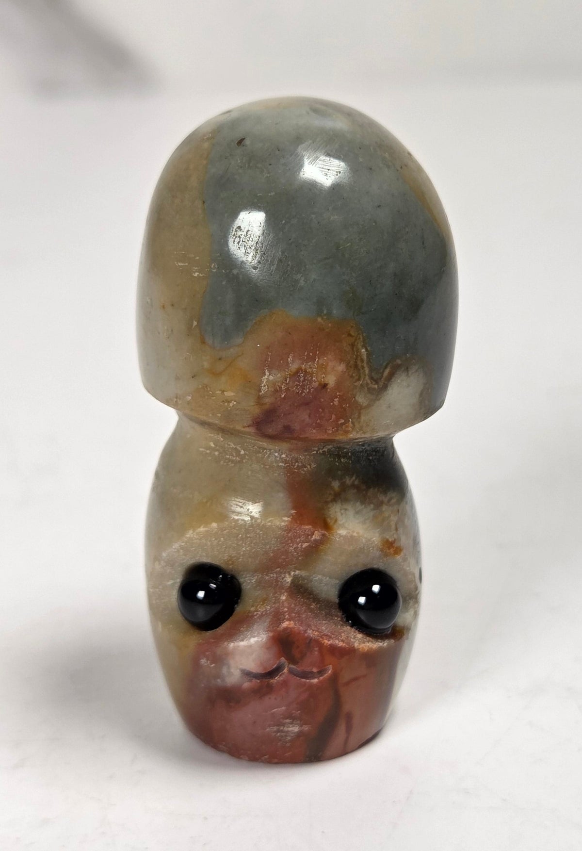 A small carved stone figurine shaped like a mushroom cap resting on a mushroom stem, featuring black bead eyes and a mottled pattern of gray, brown, and beige, set against a plain light background.