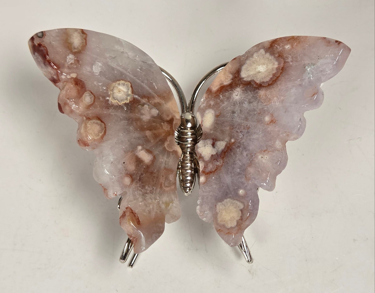 A butterfly-shaped resin brooch with textured, organic patterns in shades of pink, brown, and cream, attached to a silver-tone metal wire frame.