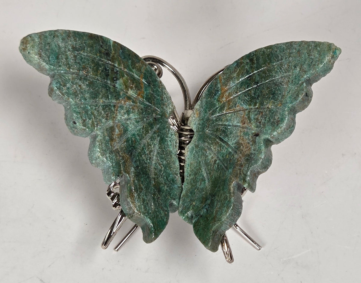 A butterfly-shaped brooch featuring textured green stone wings with brown marbling, secured by silver-tone metal rings and pins on a light gray background.