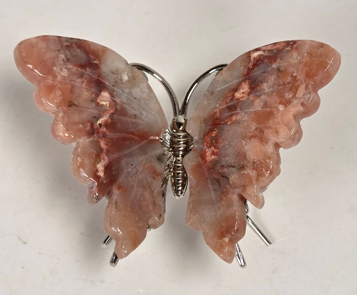 A butterfly-shaped brooch featuring carved pinkish-brown stone wings with silver wire wrapping at the body and legs, shown on a plain off-white background.