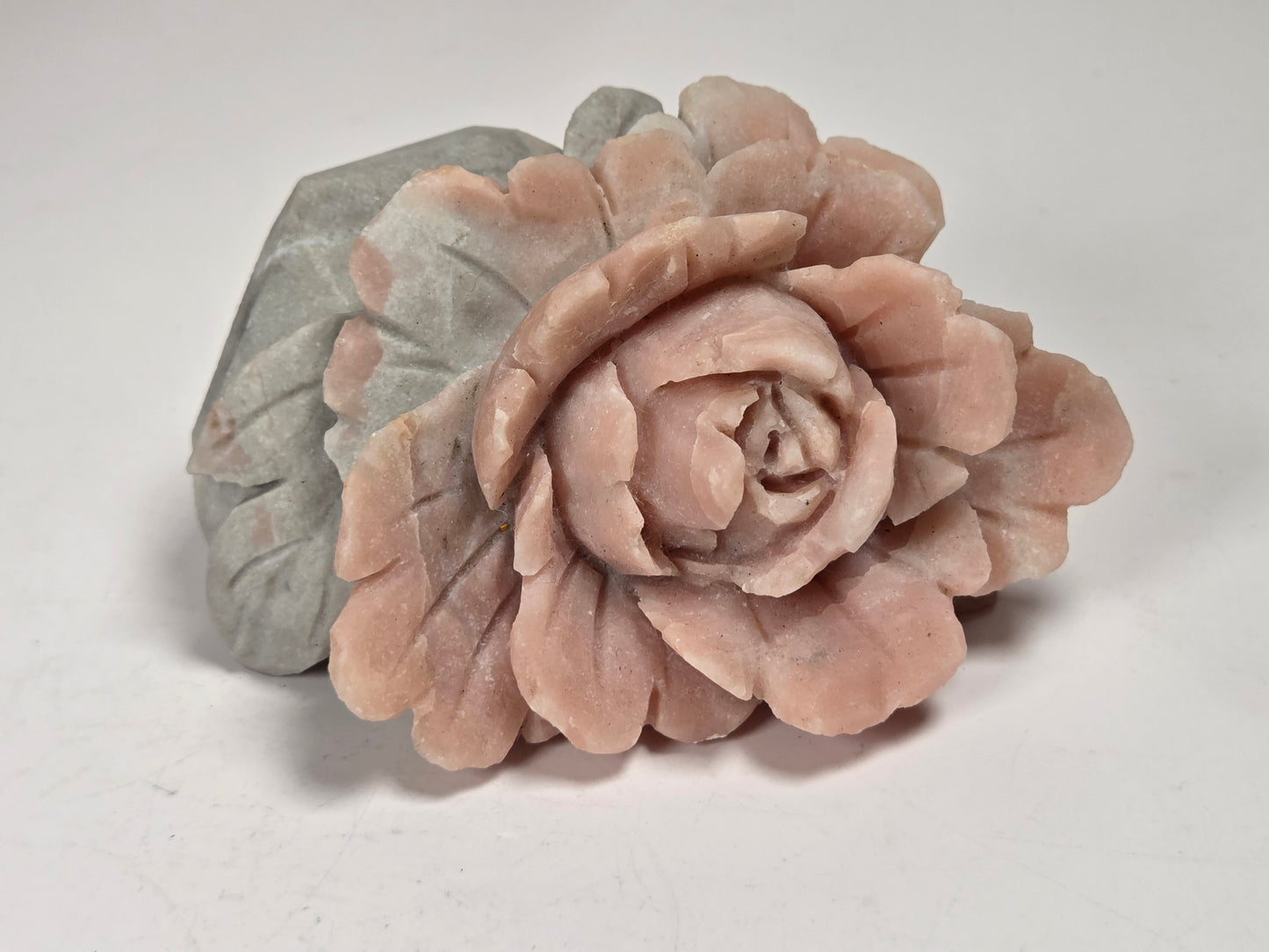 A carved stone rose figurine in a dusty pink color, positioned in front of a matching gray stone leaf base on a plain light background.