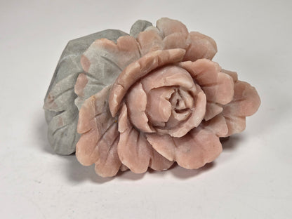 A carved stone rose figurine in a dusty pink color, positioned in front of a matching gray stone leaf base on a plain light background.