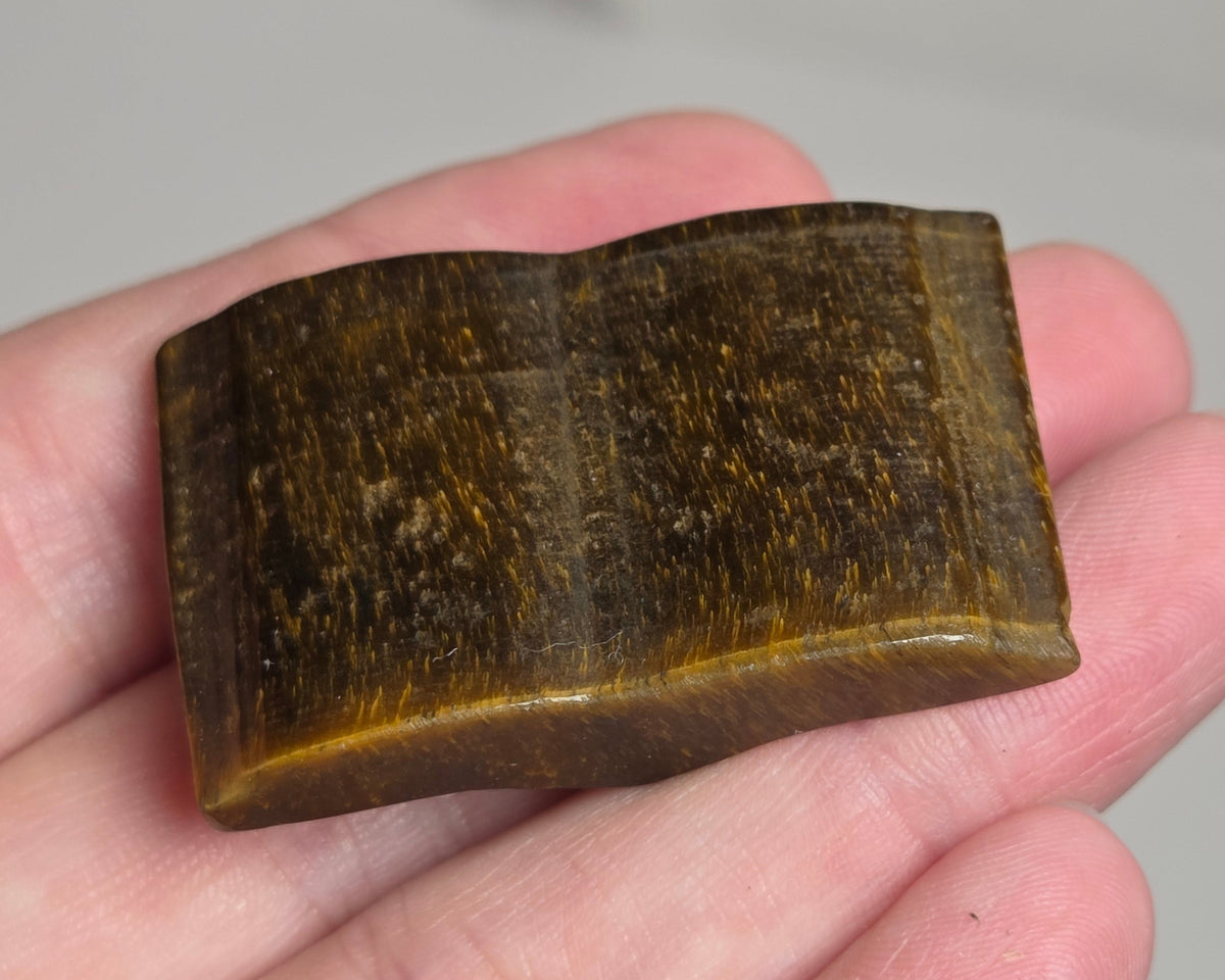 A small polished brown stone or wooden block with a curved rectangular shape, held in a person's hand against a light gray background.