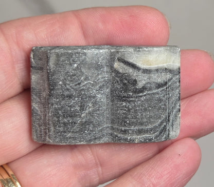 A hand holding a small rectangular gray stone or polished rock slab with natural marbled patterns, against a light gray marble-like background.