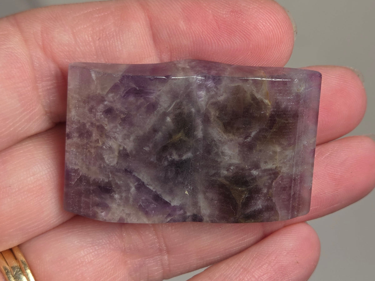 A hand holding a polished rectangular purple stone with natural marbled patterns, shown against a soft gray background.