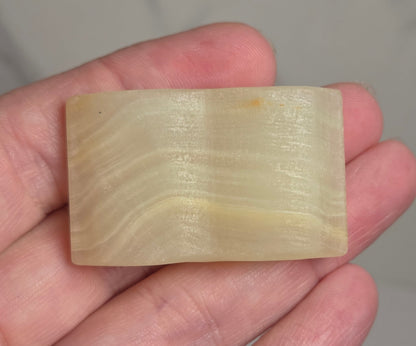 A hand holding a small rectangular pale yellow stone or resin block with subtle horizontal striations, against a light gray marble-like background.