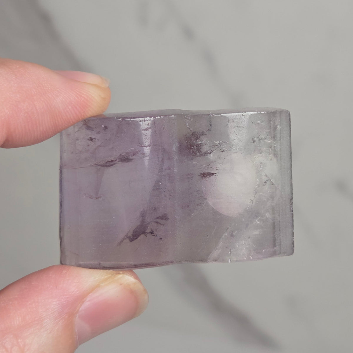 A hand holding a small, semi-translucent lavender-purple crystal or gemstone with natural inclusions, set against a light gray marble background.