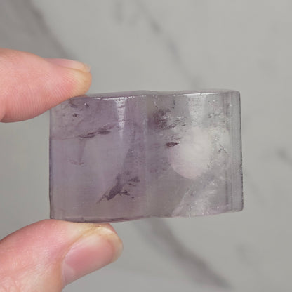 A hand holding a small, semi-translucent lavender-purple crystal or gemstone with natural inclusions, set against a light gray marble background.