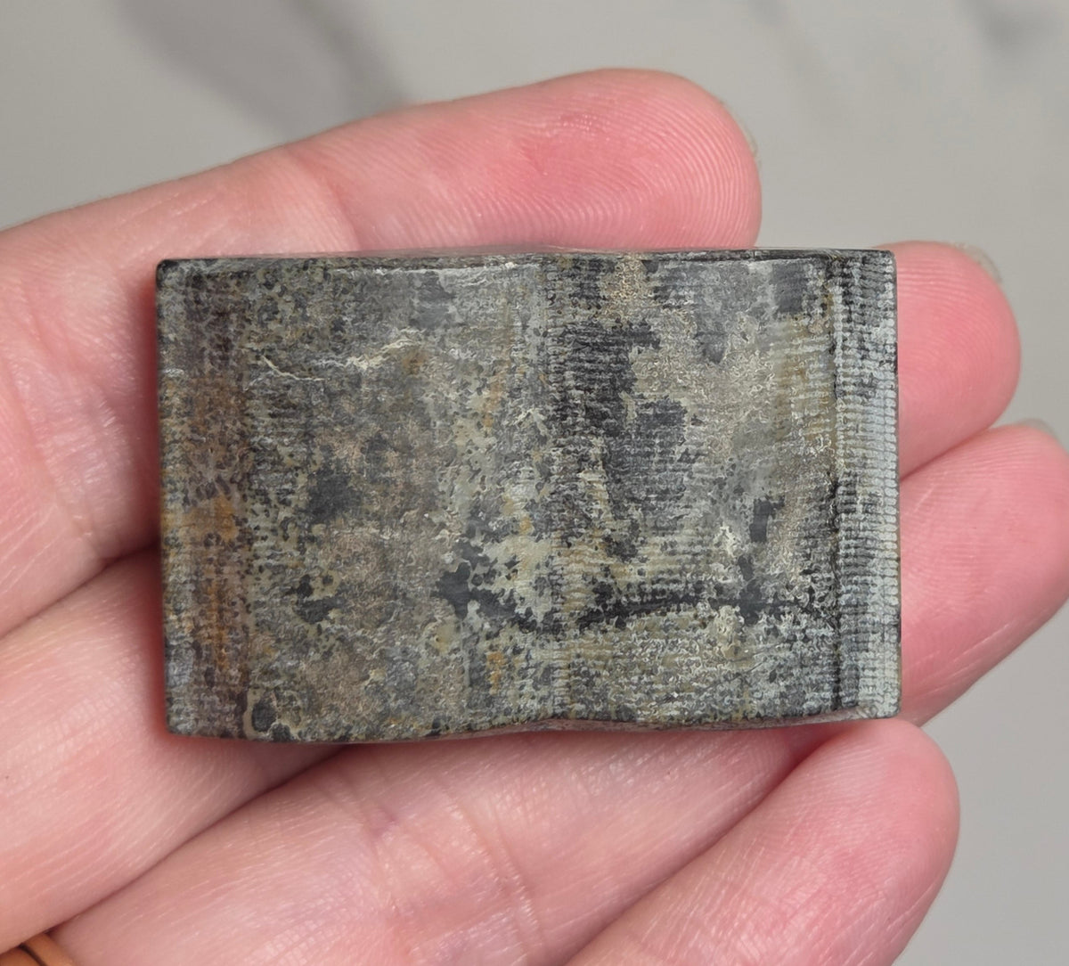 A rectangular textured gray stone or ceramic tile held in a person's hand, with a gold ring visible on the ring finger against a light gray marble-patterned background.