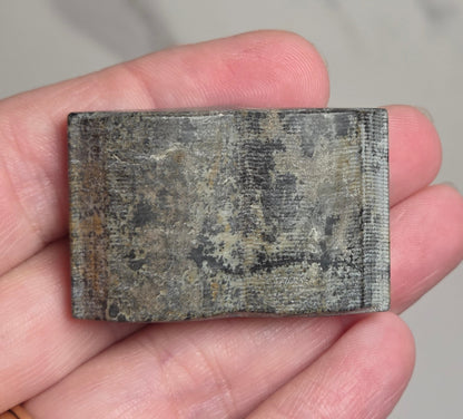 A rectangular textured gray stone or ceramic tile held in a person's hand, with a gold ring visible on the ring finger against a light gray marble-patterned background.