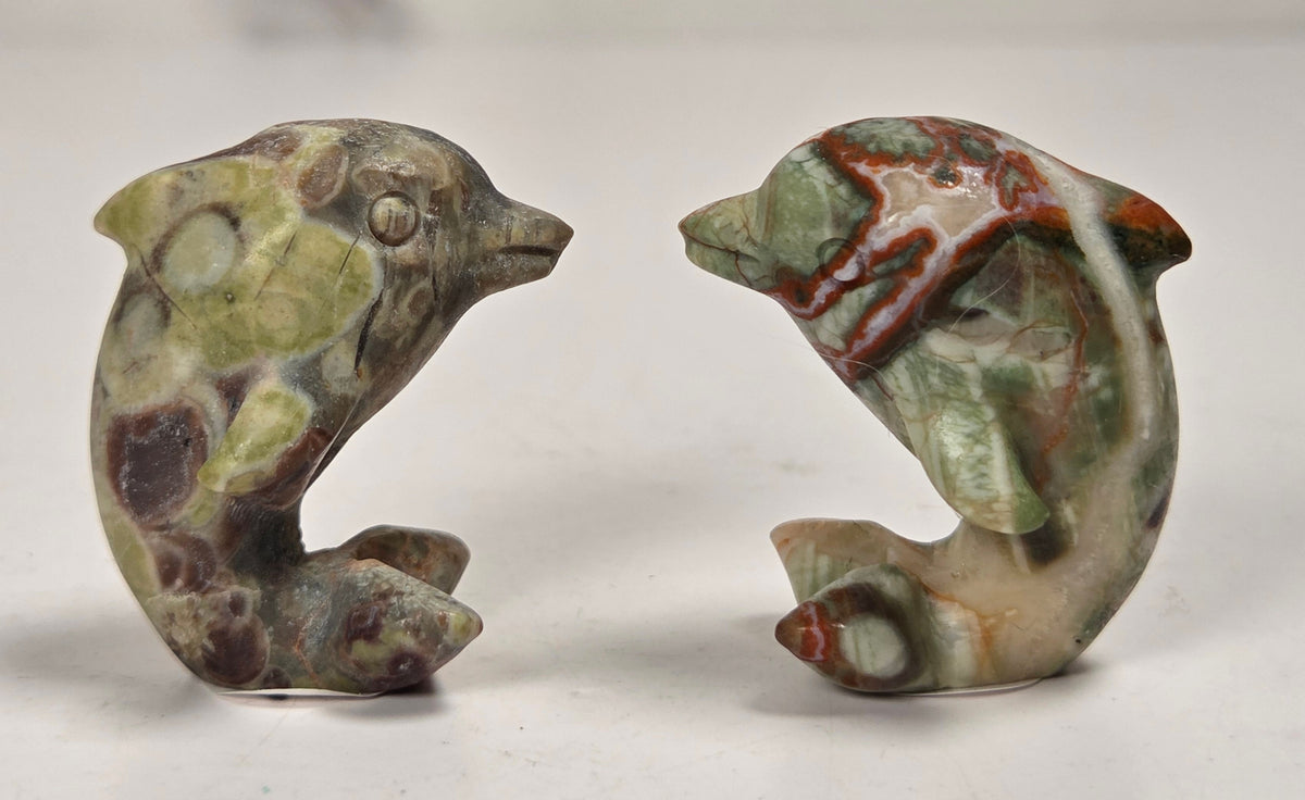 A pair of small carved stone dolphin figurines with a natural marbled pattern in green, brown, beige, and reddish tones, displayed on a light surface.