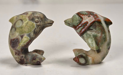 A pair of small carved stone dolphin figurines with a natural marbled pattern in green, brown, beige, and reddish tones, displayed on a light surface.