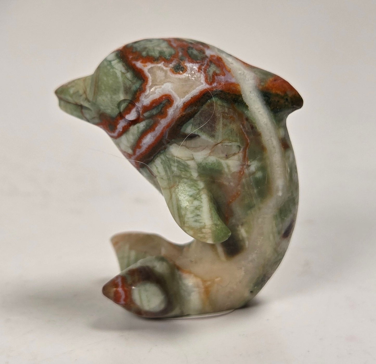 A small carved figurine of a dolphin or whale made from marbled stone with green, beige, and reddish-brown patterns, standing on a light gray surface.