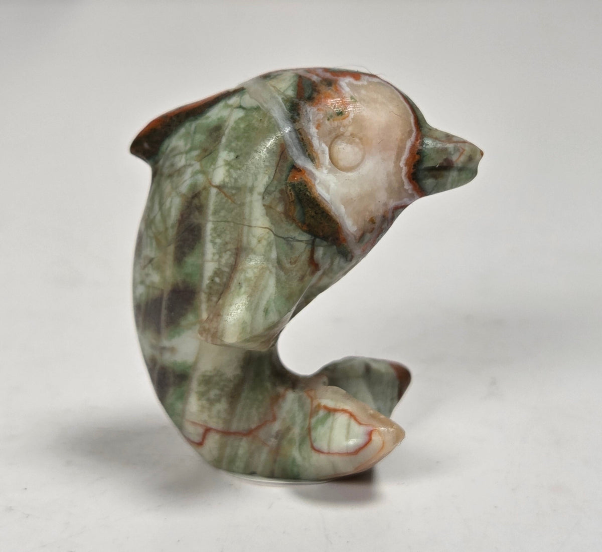 A small carved stone figurine shaped like a dolphin or whale, featuring natural marbled patterns in green, beige, brown, and reddish tones, photographed on a plain light background.