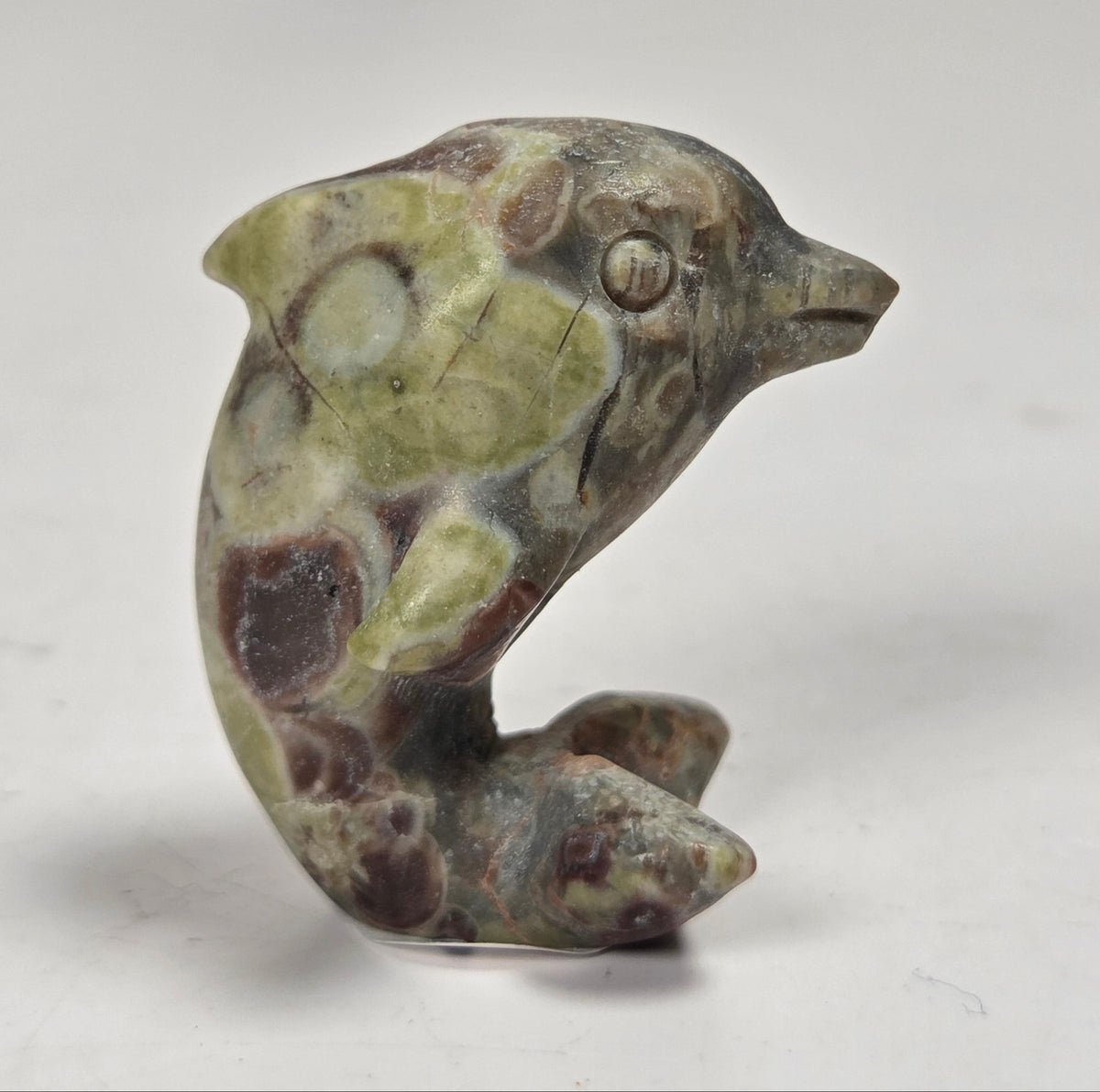 A small carved stone dolphin figurine with a mottled pattern of olive green, brown, and dark reddish-brown, standing on a plain light gray surface.