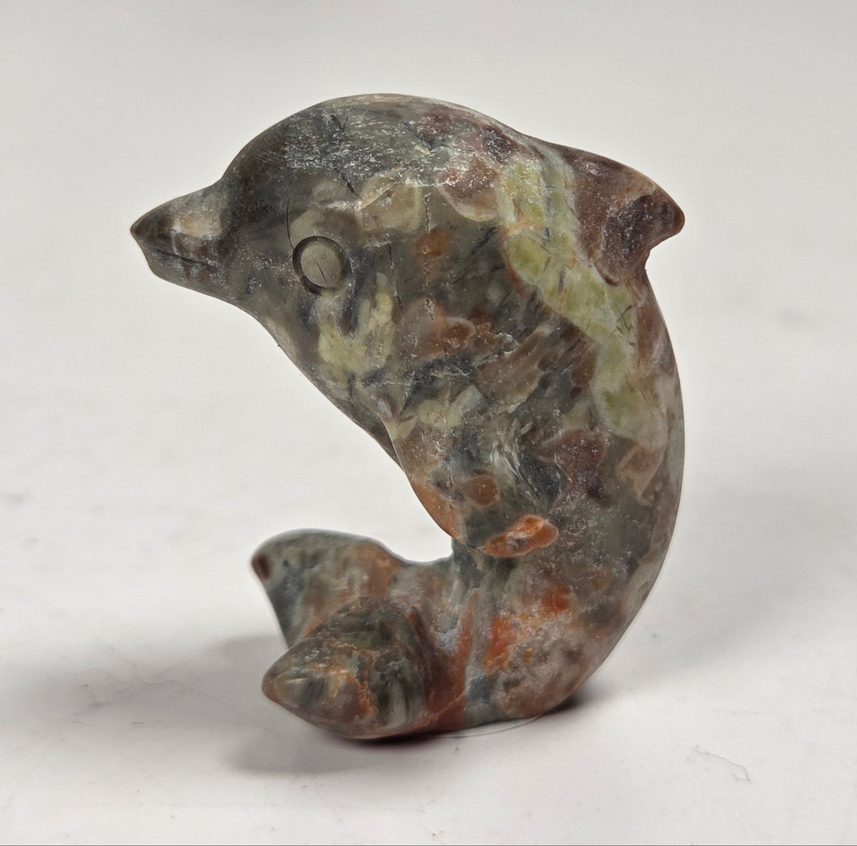 A small carved stone dolphin figurine with natural marbled textures in gray, green, brown, and rust tones, photographed on a plain light background.