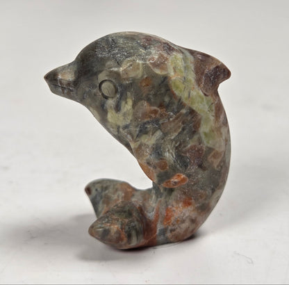 A small carved stone dolphin figurine with natural marbled textures in gray, green, brown, and rust tones, photographed on a plain light background.
