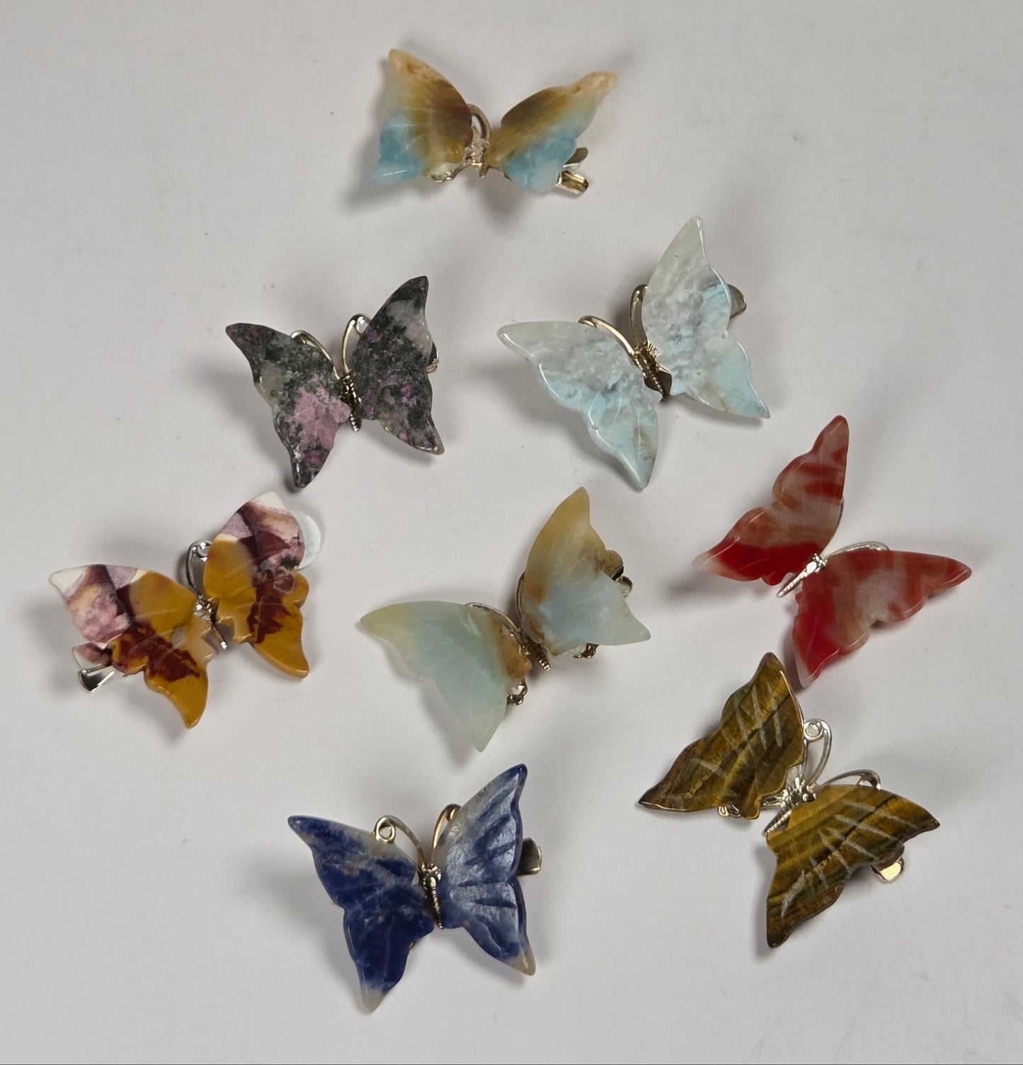 Nine butterfly-shaped earrings made from colorful stone inlay, arranged on a plain off-white background.