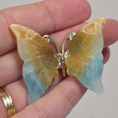 A butterfly-shaped pendant or brooch made from marbled stone with yellow, blue, and white tones, held on a person's fingers. The butterfly has a gold-tone metal body and wings.